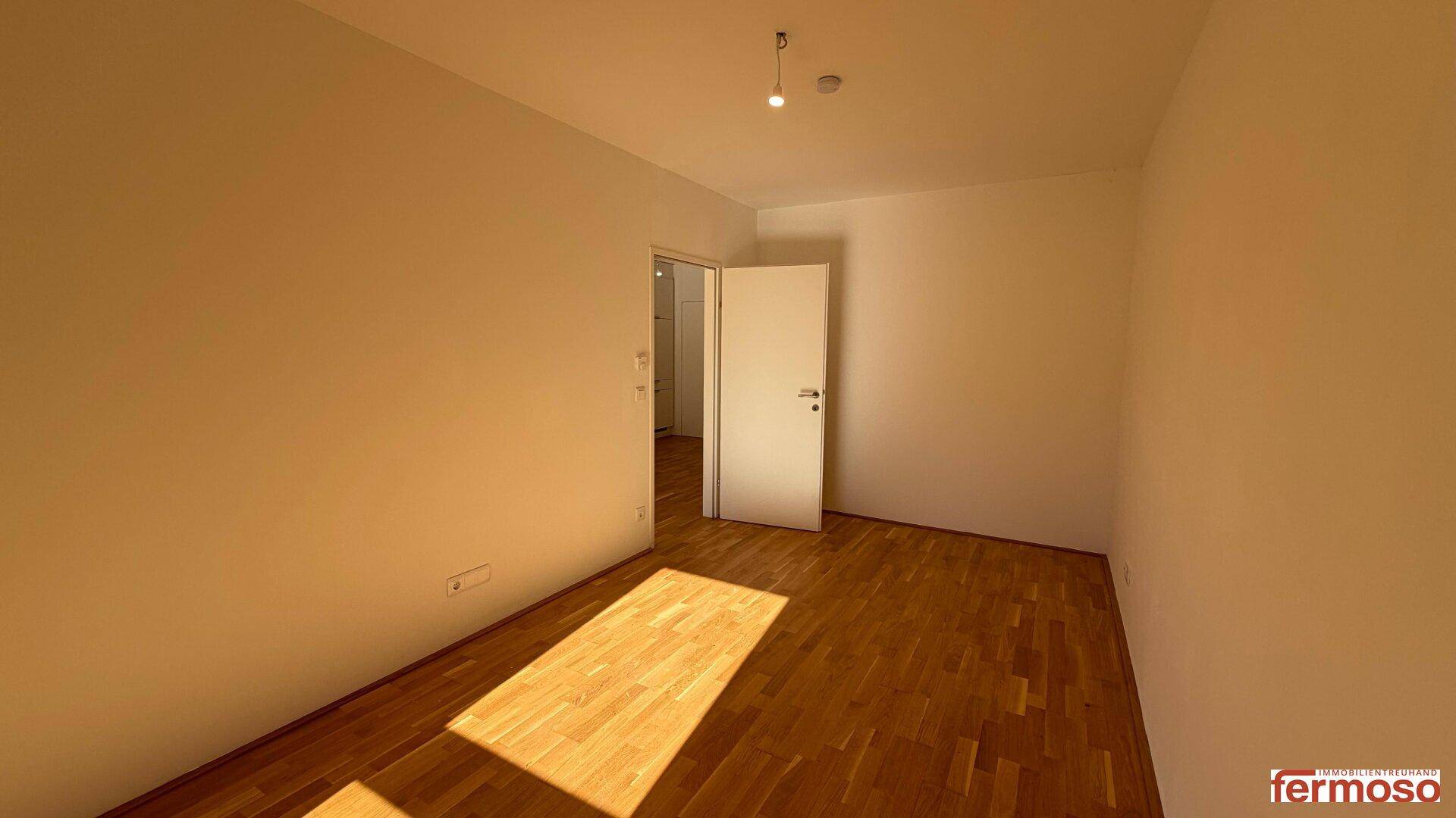 Sonniger, leerer Raum mit Parkettboden und einer Tür, die zu einem weiteren Zimmer führt.