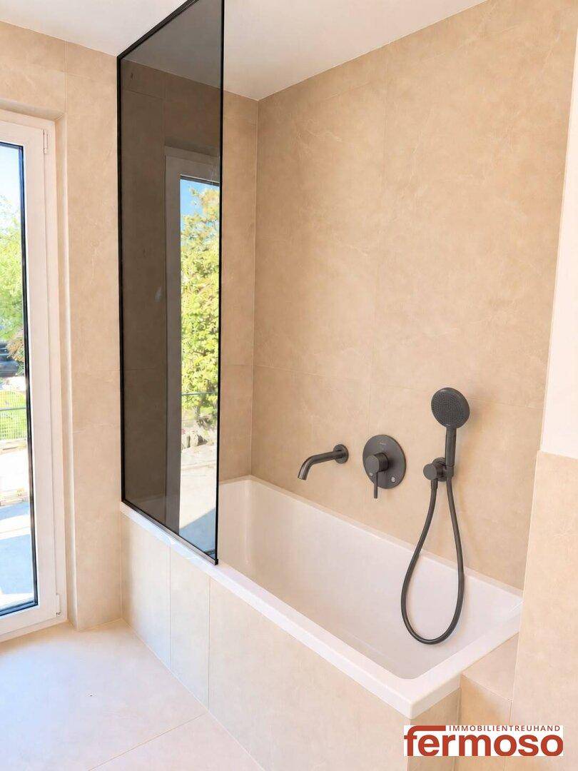 Helles Badezimmer mit Badewanne, Glasduschwand und Fenster, ausgestattet mit beigefarbenen Fliesen.