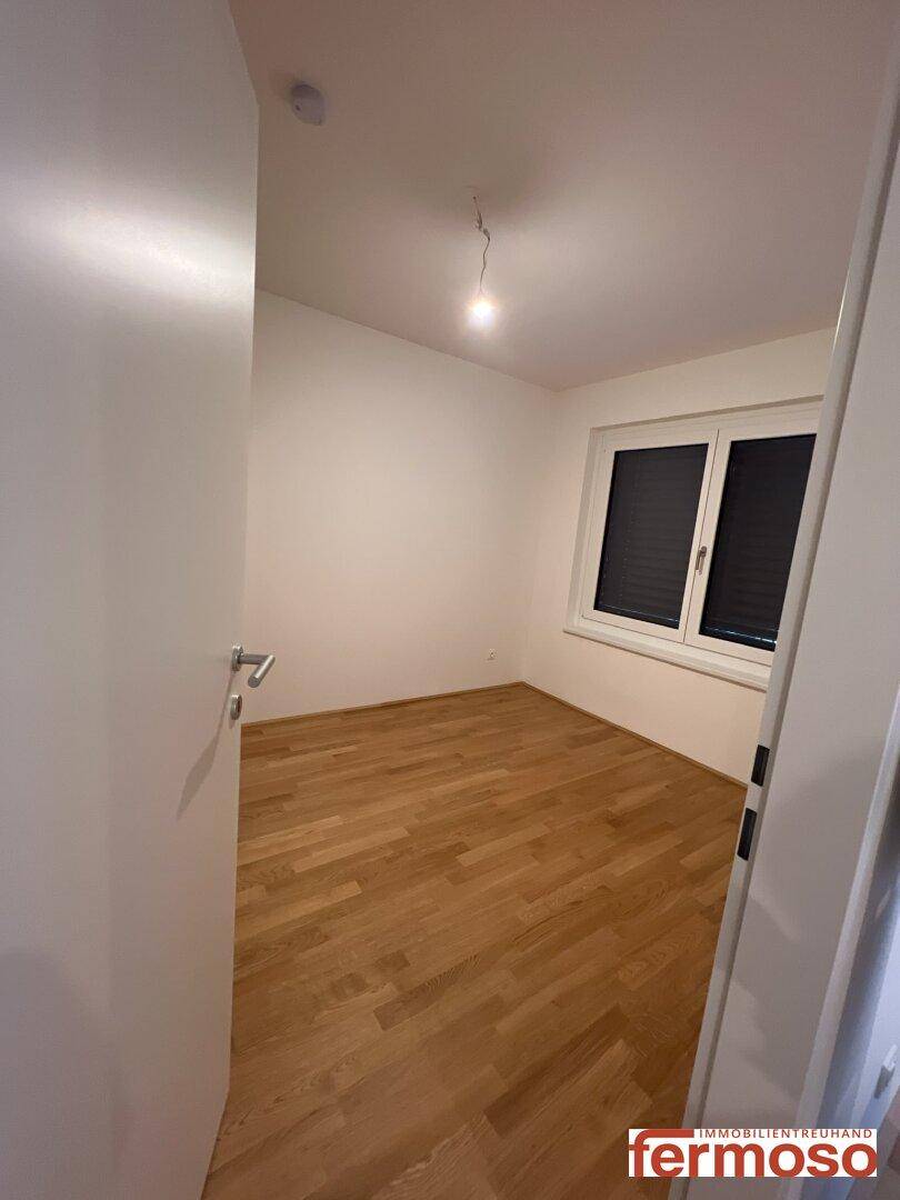 Leeres Schlafzimmer mit hellem Parkettboden und Fenster mit Jalousien.
