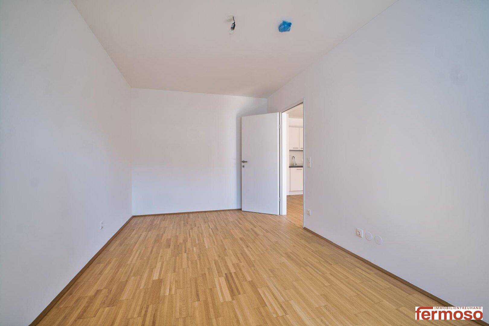 Leeres Zimmer mit hellem Parkettboden und Zugang zu einem weiteren Raum.