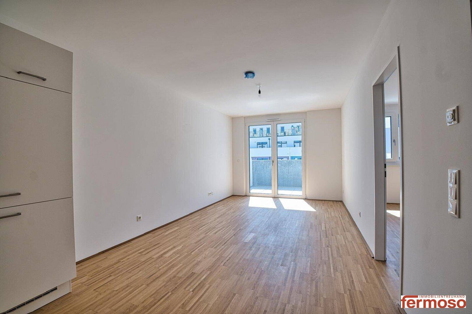 Helles Zimmer mit Parkettboden und Zugang zu einem Balkon, viel Tageslicht.