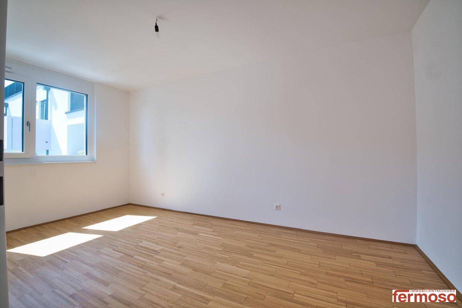 Leeres Schlafzimmer mit hellem Parkettboden und Fenster, das viel Licht hereinlässt.
