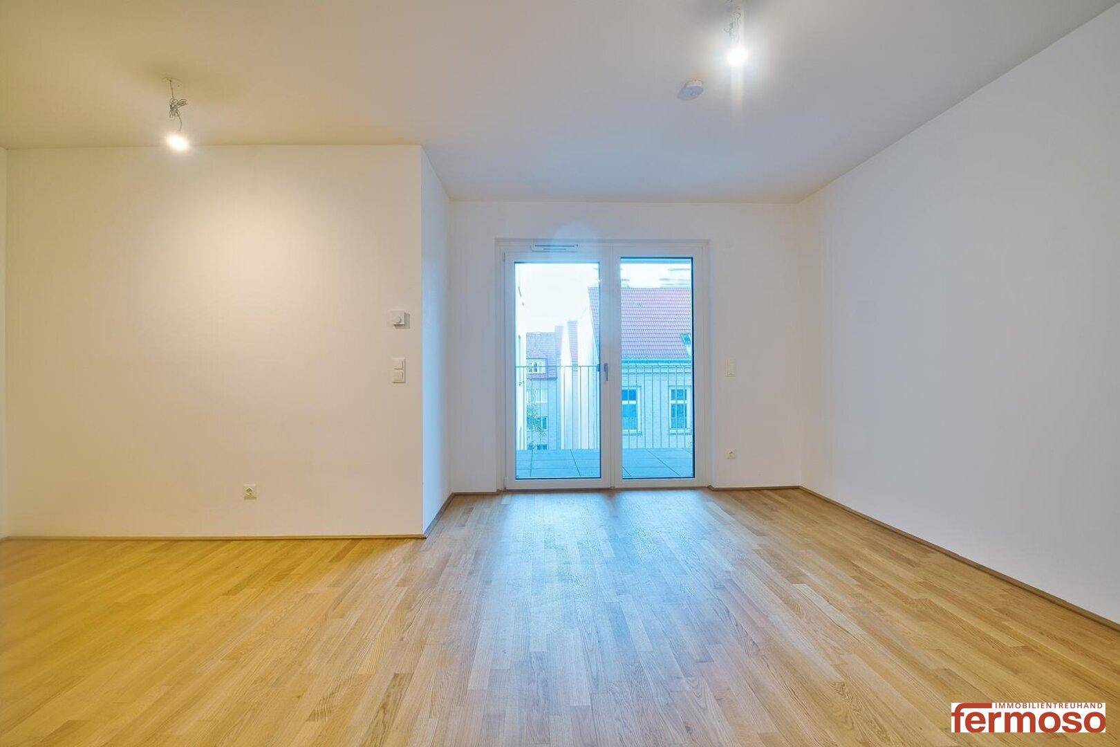 Leerer Wohnraum mit hellem Holzboden und französischem Balkon.