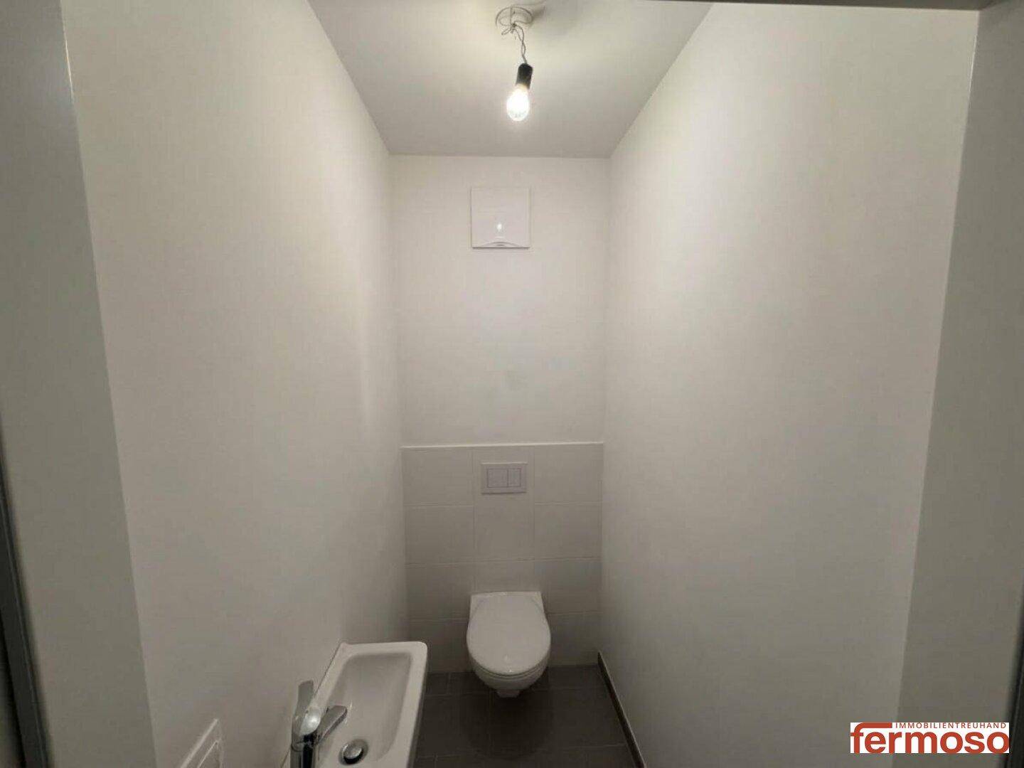 Separates WC mit wandhängender Toilette, kleinem Waschbecken und Fliesenboden.