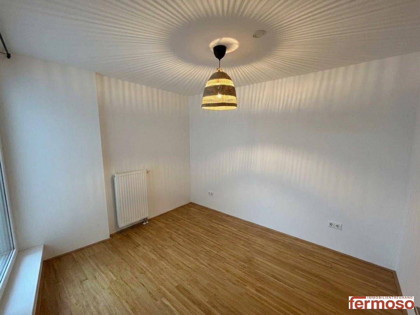 Heller, leerer Raum mit Parkettboden, Heizkörper und einem Fenster.