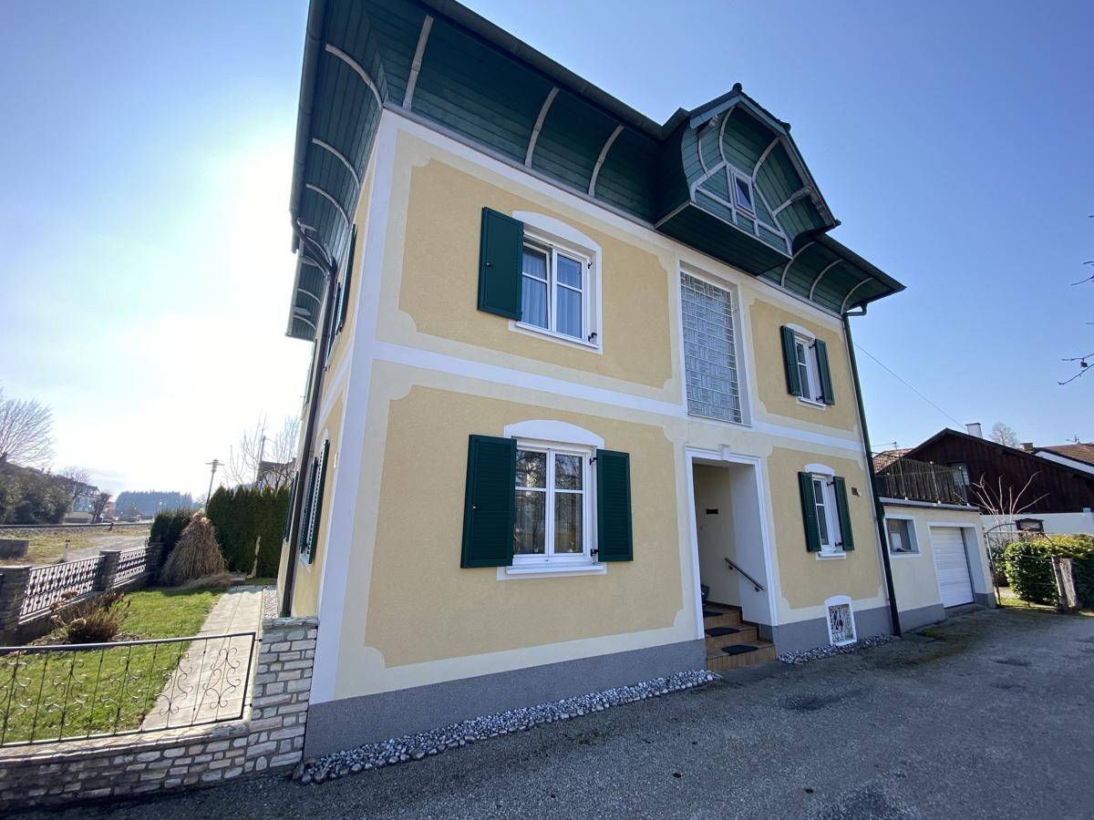 Vorderansicht des gelben Wohnhauses mit grünen Fensterläden und Eingangsbereich unter der Sonne.