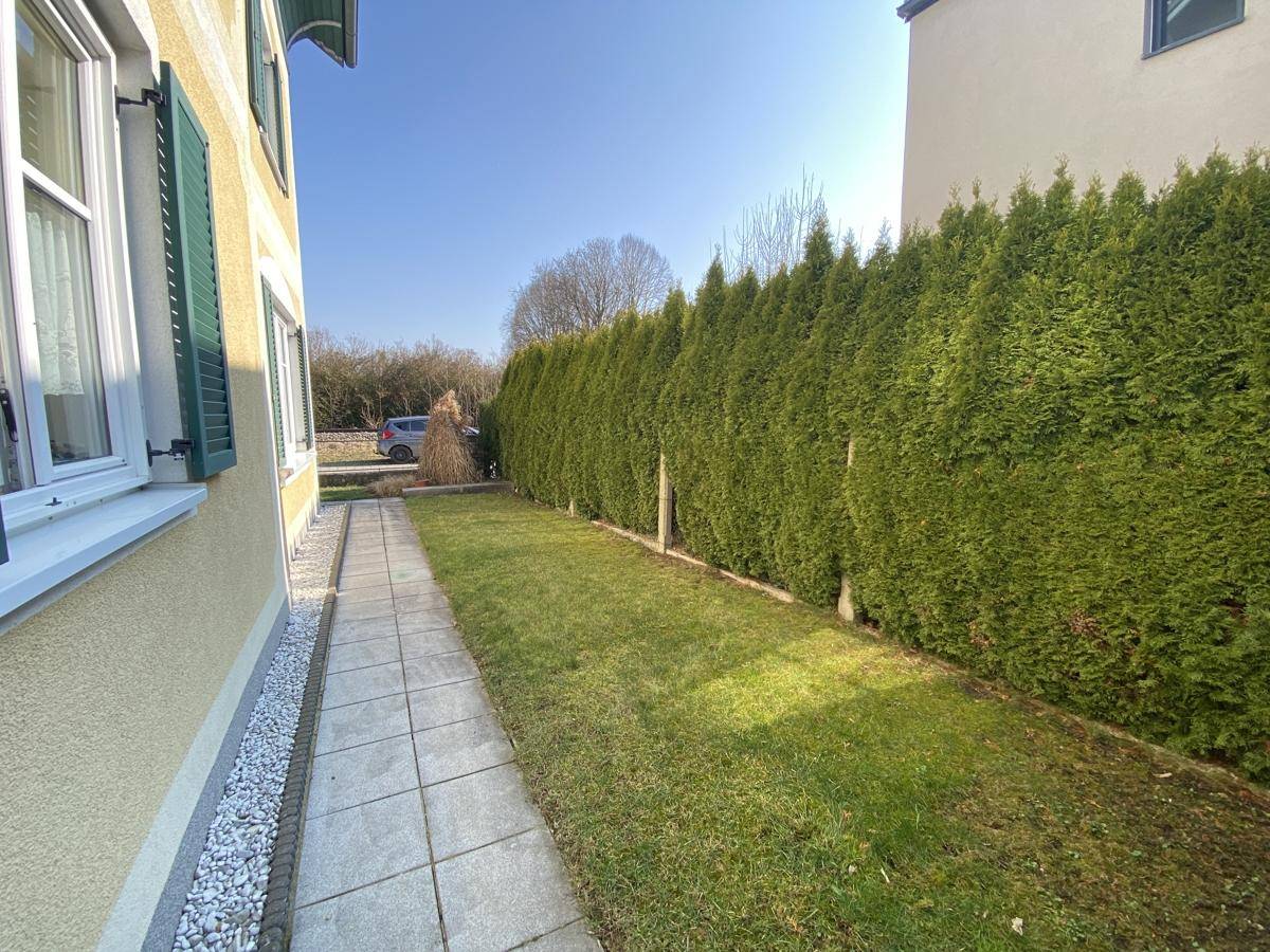 Seitlicher Gartenweg mit Rasenfläche und hoher Hecke entlang der Hauswand.