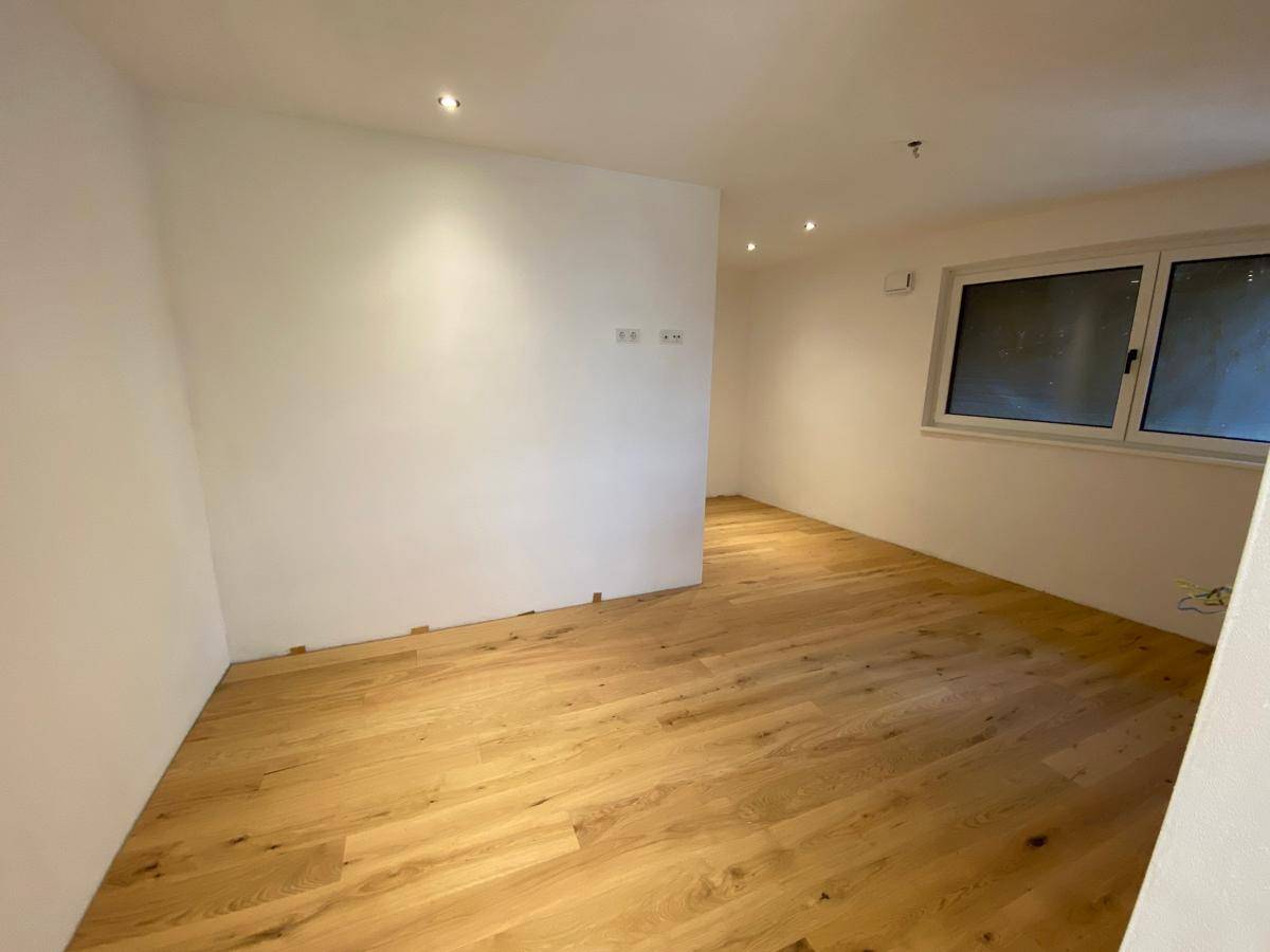 Geräumiges Zimmer mit Holzboden, weißen Wänden und integrierter Deckenbeleuchtung.