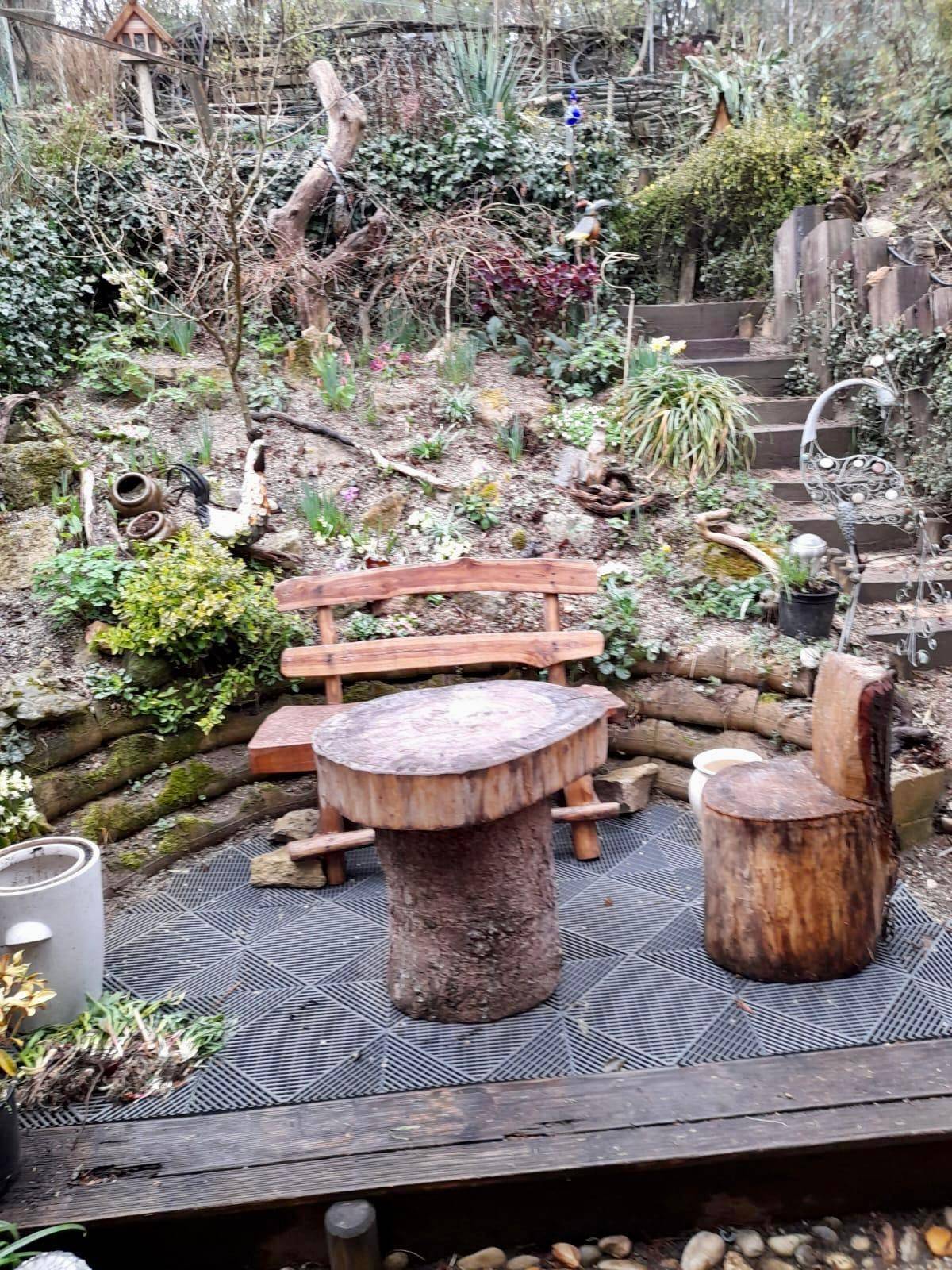 Rustikaler Sitzbereich im Garten mit Holzbänken und Tisch aus Baumstämmen, umgeben von Pflanzen.