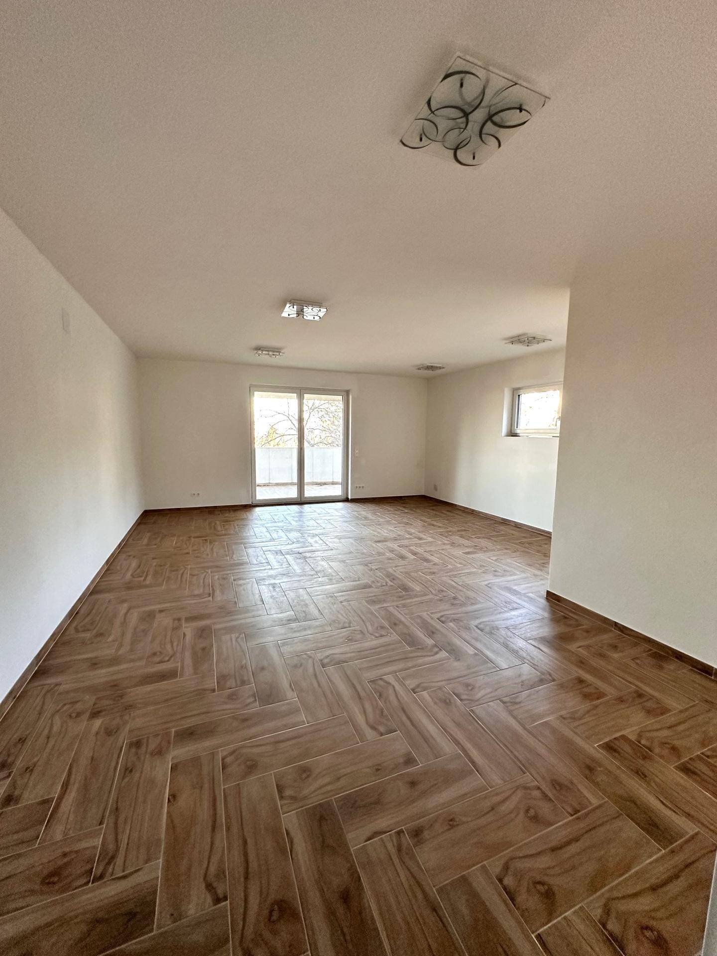 Geräumiger, heller Raum mit dekorativem Fliesenboden in Holzoptik und Balkonzugang.