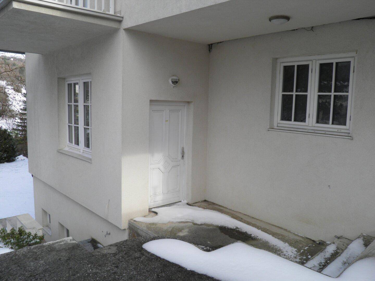 Eingangsbereich des Hauses mit weißer Tür und Fenstern, umgeben von winterlichem Schnee.
