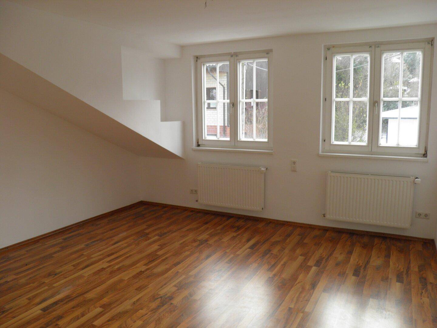 Helles Zimmer mit Holzboden, zwei Fenstern und einer Dachschräge an der Seite.