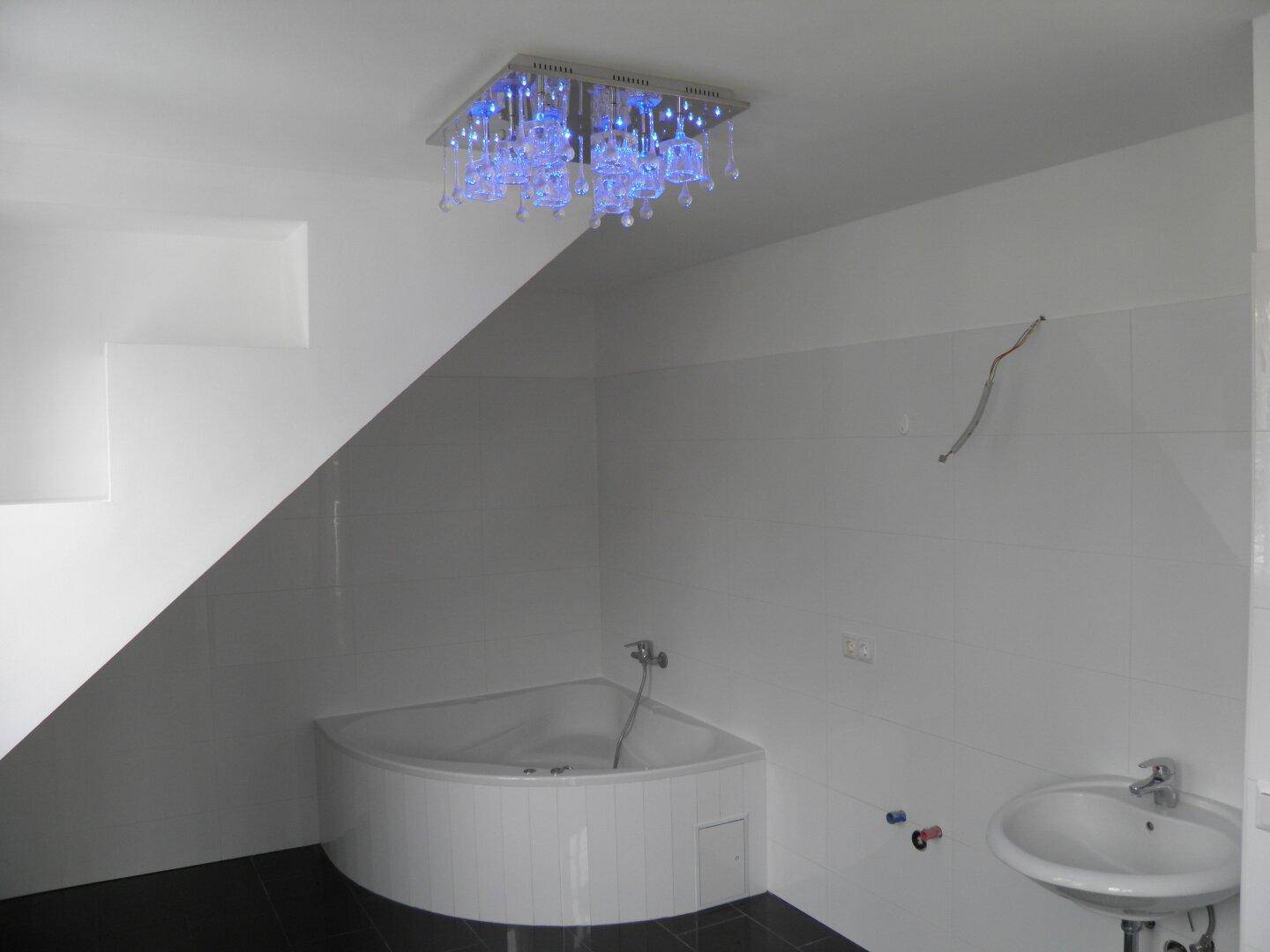 Modernes Badezimmer mit weißer Eckbadewanne, Waschbecken und blauer Deckenbeleuchtung unter der Dachschräge.