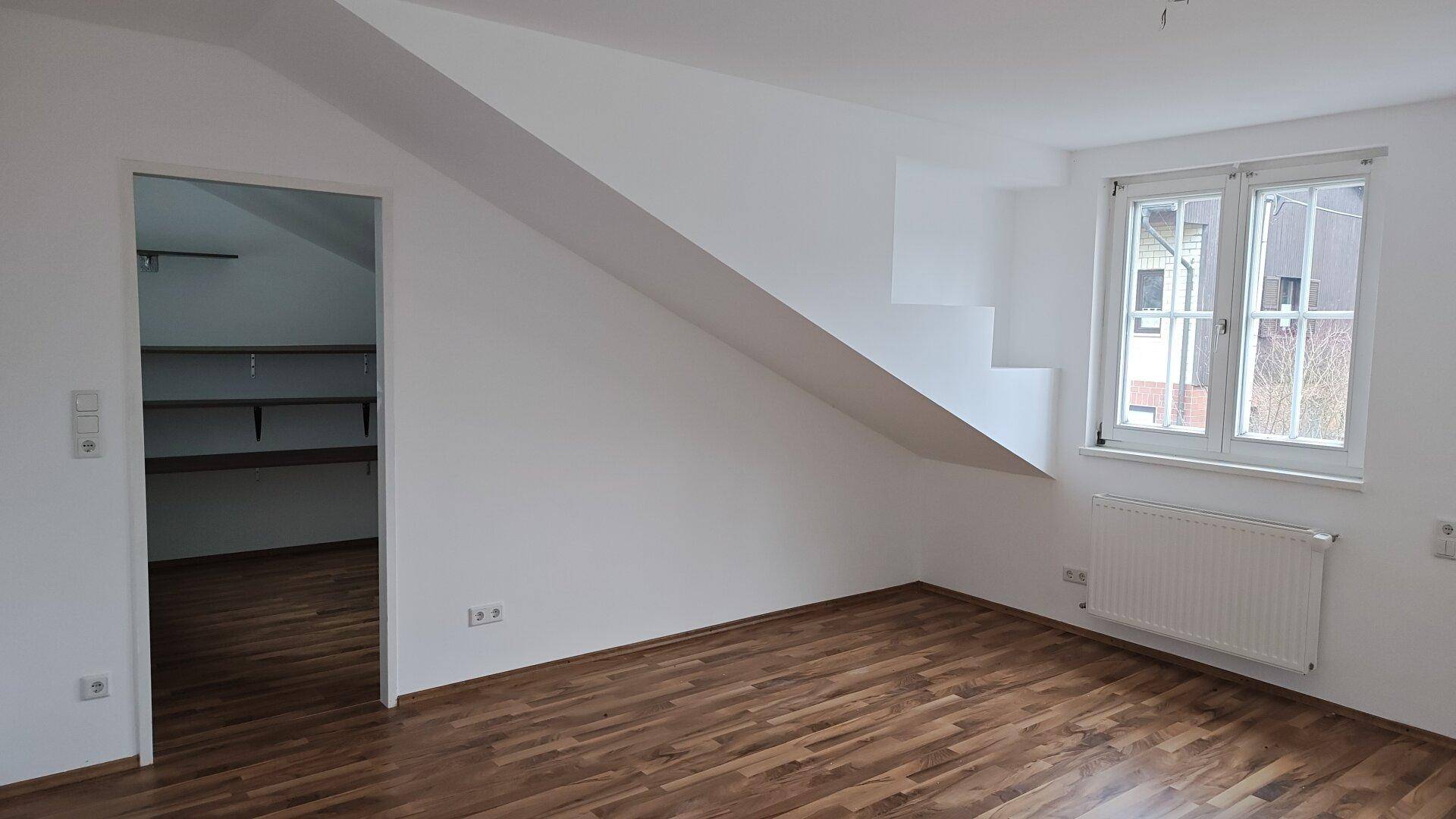 Geräumiges Zimmer mit Holzboden, Dachschräge und Zugang zu einem angrenzenden Abstellraum.