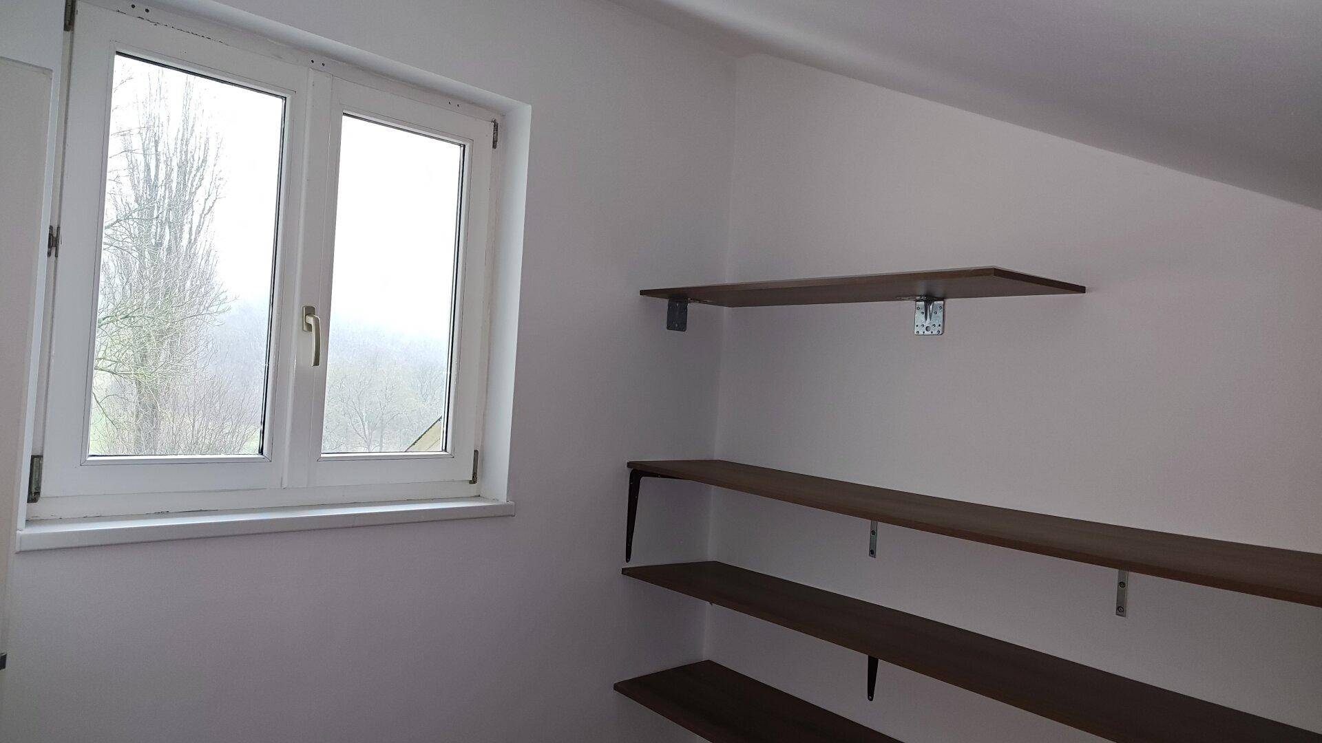 Helles Dachzimmer mit schräger Decke, Fenster und einfachen Wandregalen, ideal als Abstellraum.