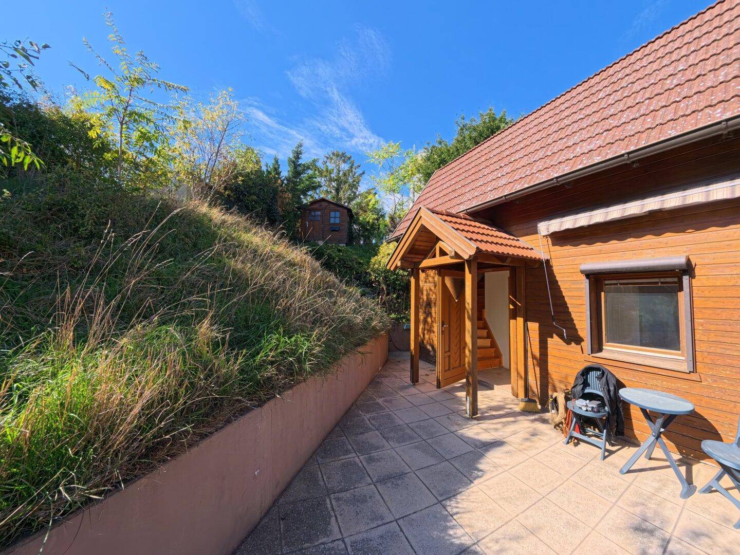 Gemütliche gepflasterte Terrasse am Hauseingang mit Sitzgelegenheiten und Blick auf den bewachsenen Hang.