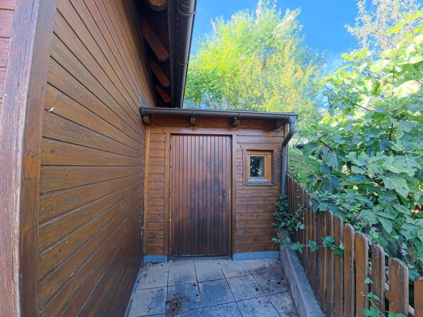 Kleiner Holzschuppen oder Anbau mit dunkler Holztür und kleinem Fenster, seitlich am Haus gelegen.
