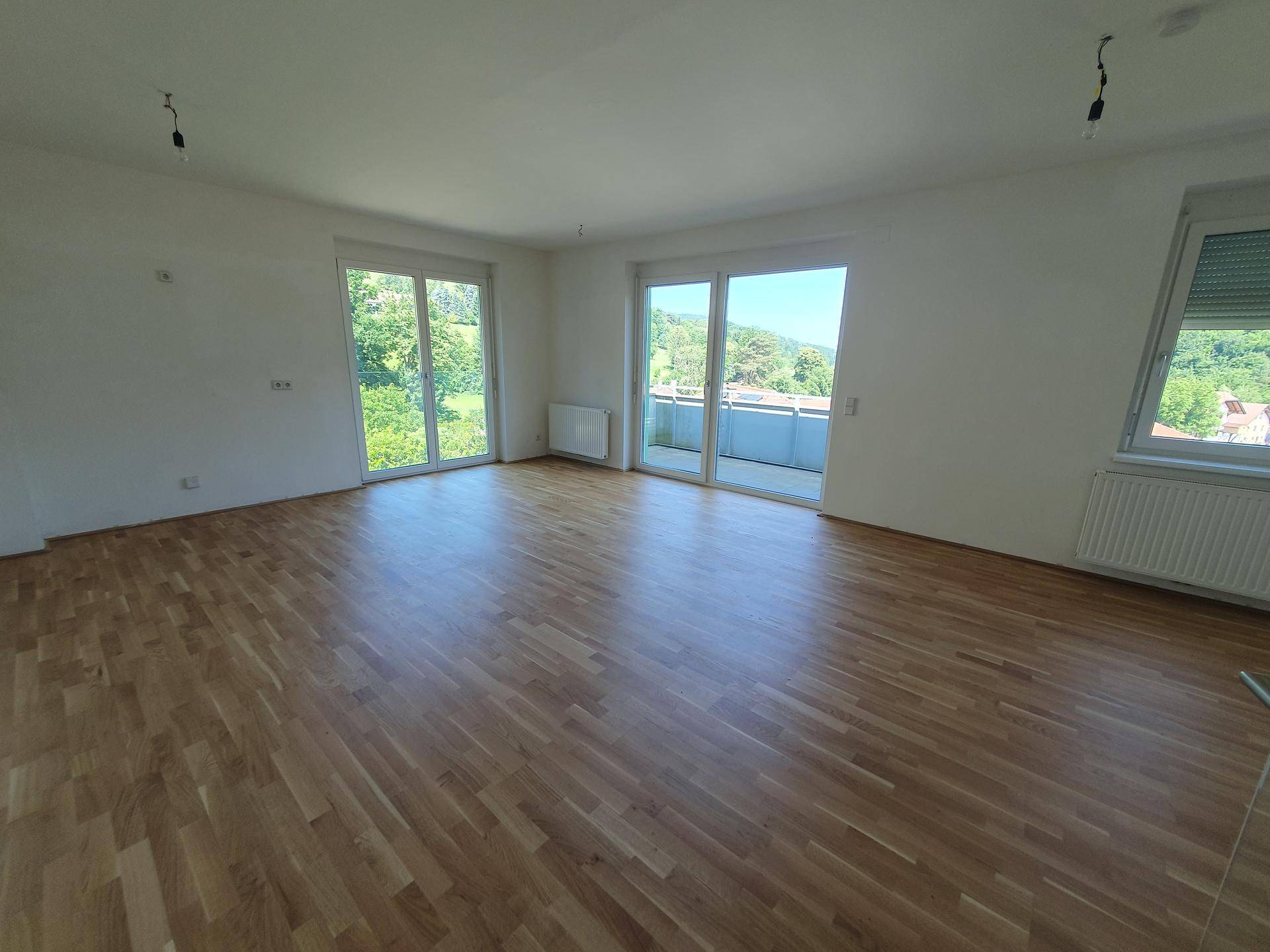 Helles, leeres Wohnzimmer mit Parkettboden, großen Fenstern und Zugang zum Balkon.