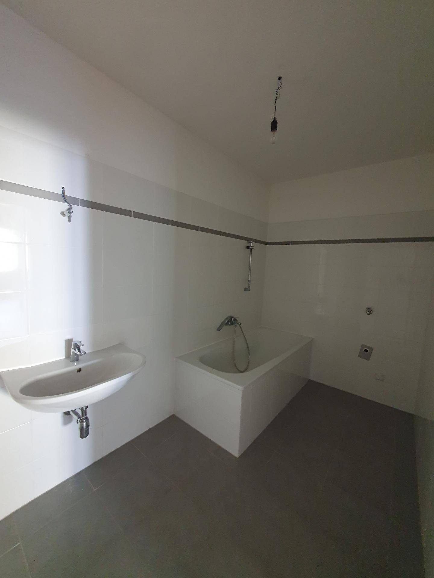 Funktionales Badezimmer mit weißen Fliesen, grauen Bodenfliesen und einer Badewanne.