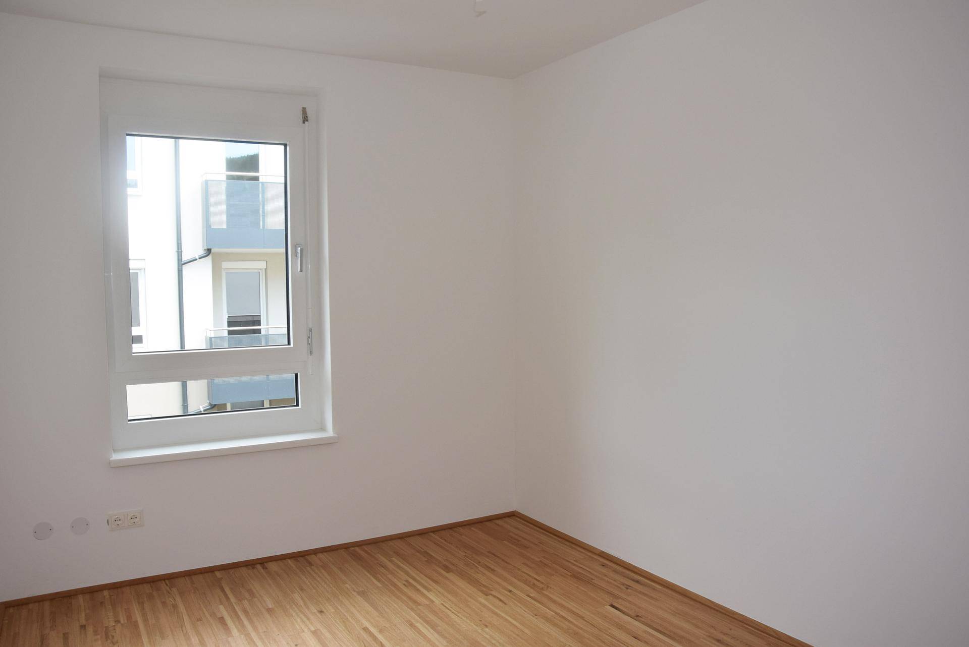 Schlichtes Zimmer mit Parkettboden und Fenster, das einen Blick nach draußen bietet.