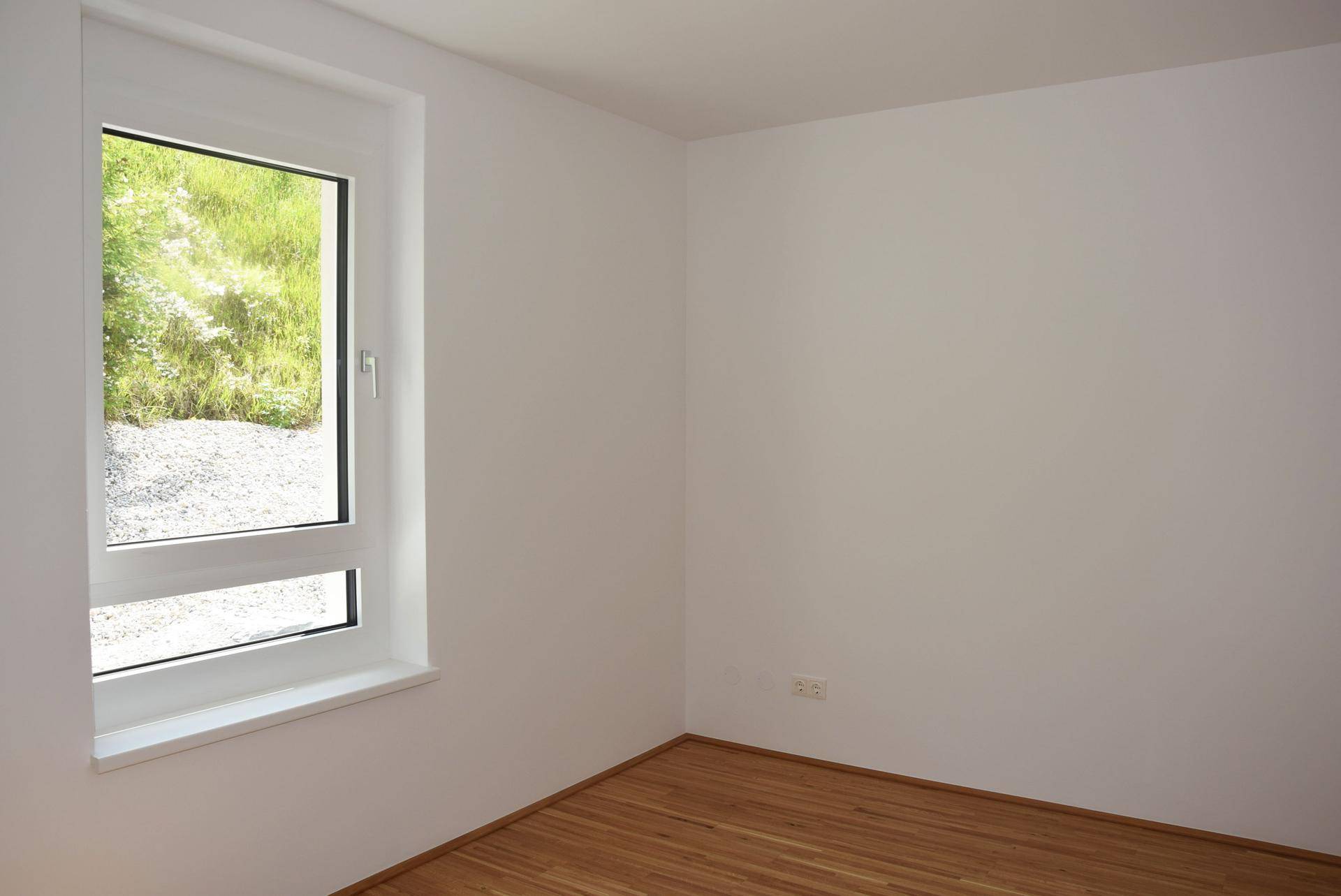 Leeres Zimmer mit Holzboden und einem großen Fenster, das viel Tageslicht hereinlässt.