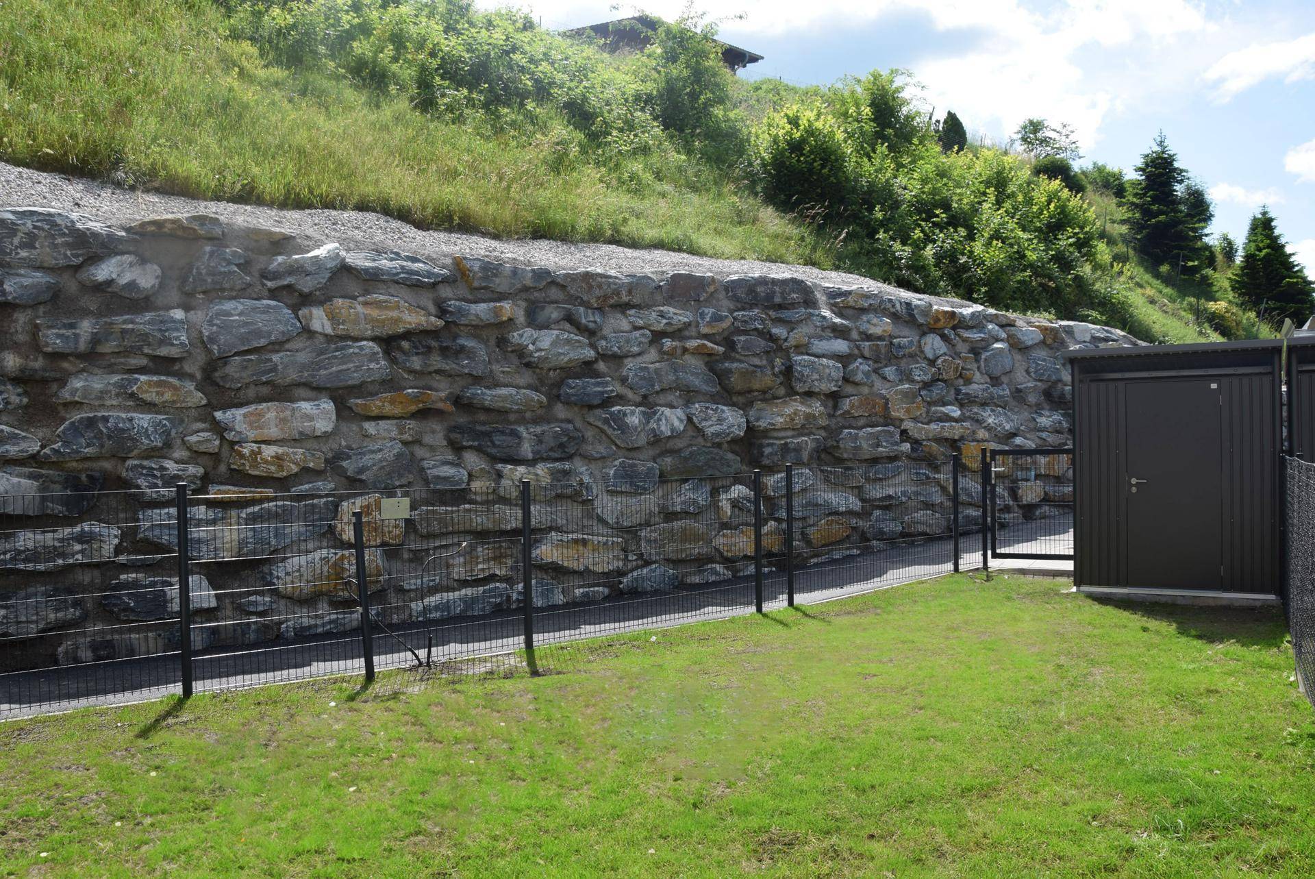 Gartenbereich mit großer Natursteinmauer, Rasenfläche und einem praktischen Geräteschuppen.