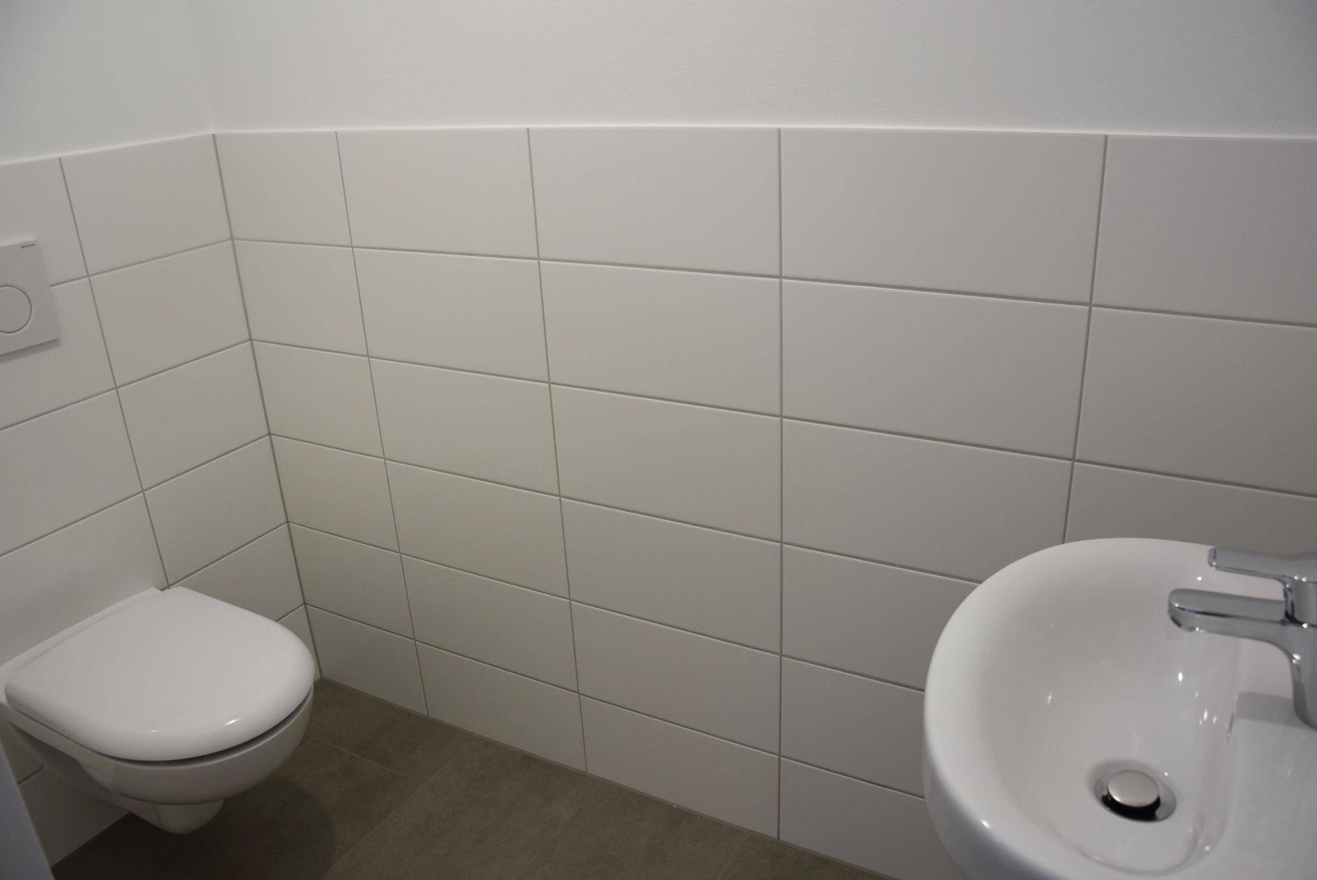 Separates WC mit wandhängender Toilette, Waschbecken und weißen Fliesen.