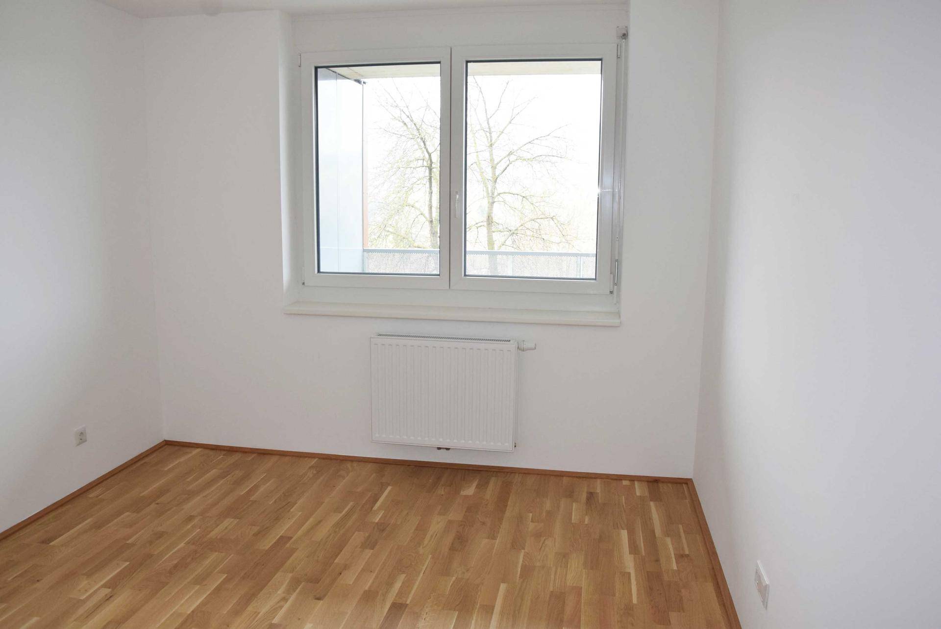 Schlafzimmer mit hellem Holzboden, weißen Wänden und einem Fenster mit Heizkörper darunter.