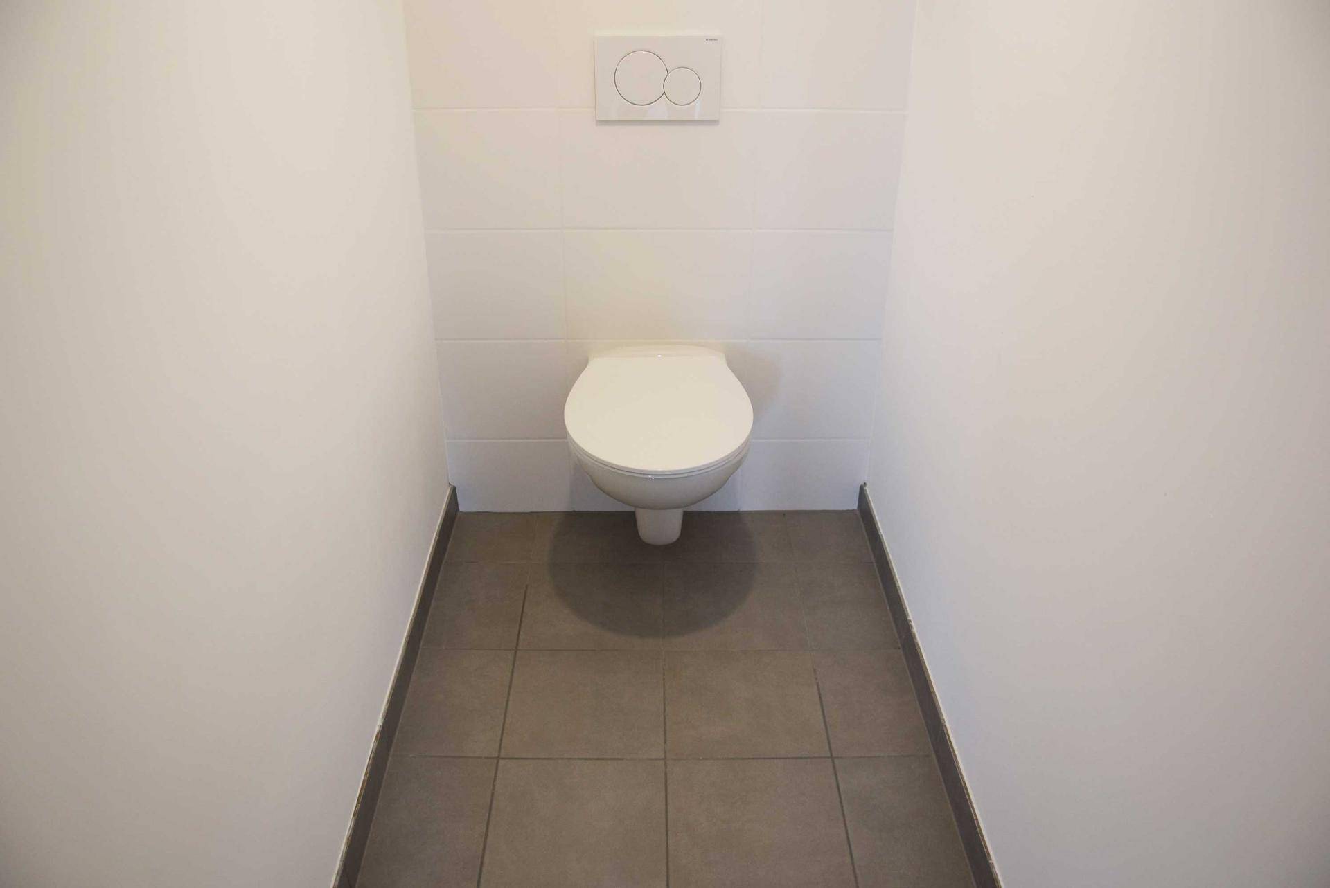 Separates WC mit wandhängender Toilette, weißen Fliesen und grauem Bodenbelag.