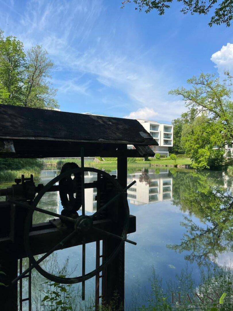 Malerischer Blick auf einen ruhigen See mit historischem Wasserrad und moderner Architektur im Hintergrund.