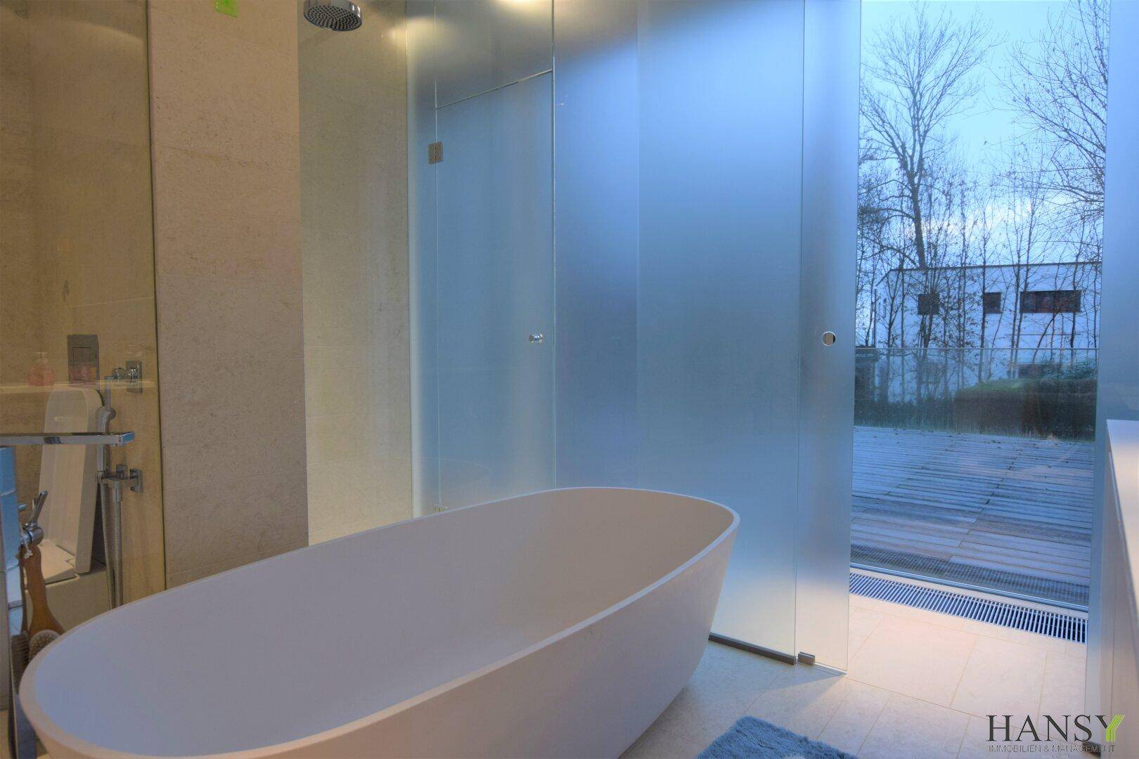 Modernes Badezimmer mit freistehender Badewanne und großer Glasdusche mit Blick ins Grüne.