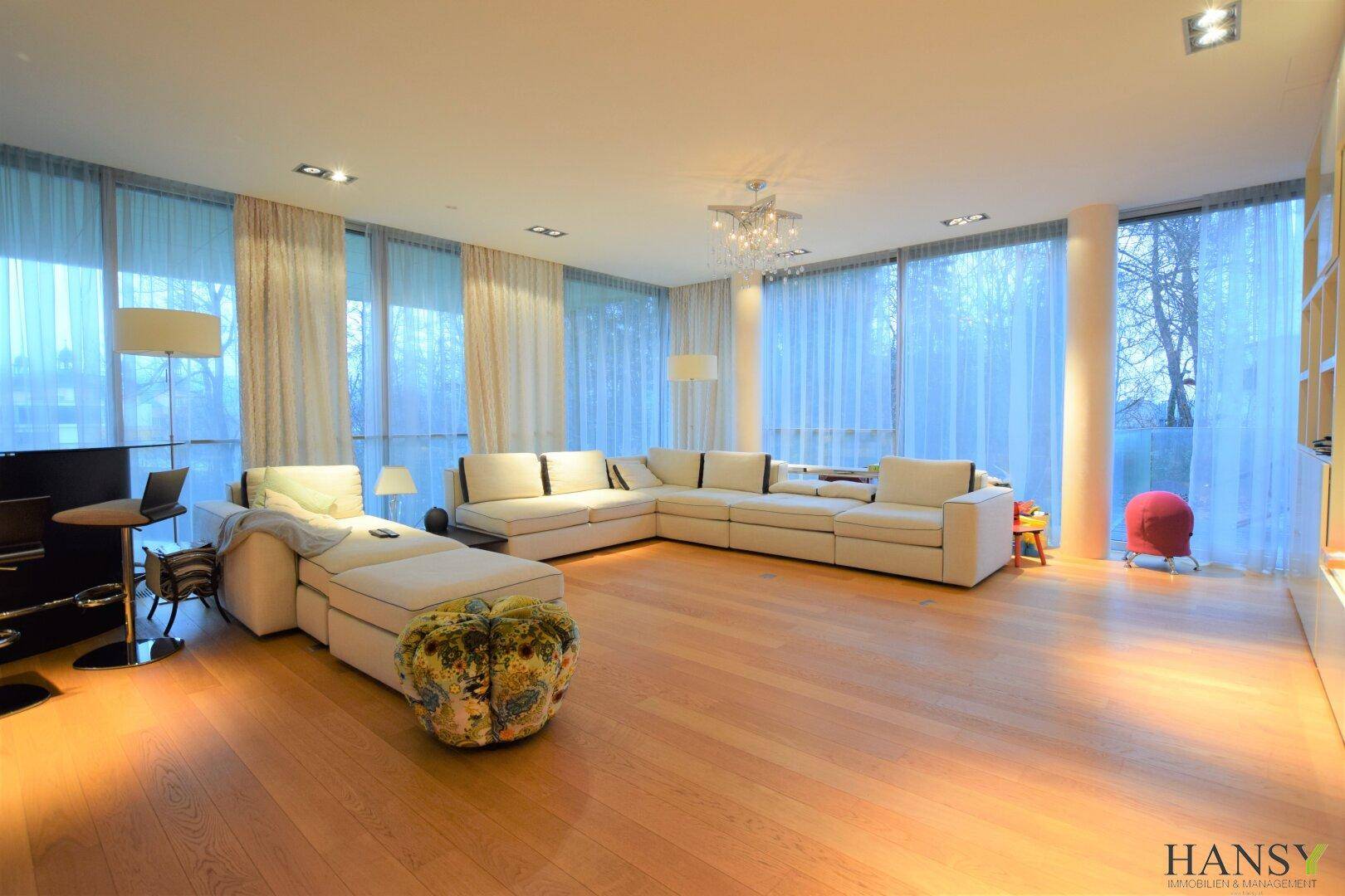 Großzügiges Wohnzimmer mit Ecksofa, Holzboden und raumhohen Fenstern für viel Licht.
