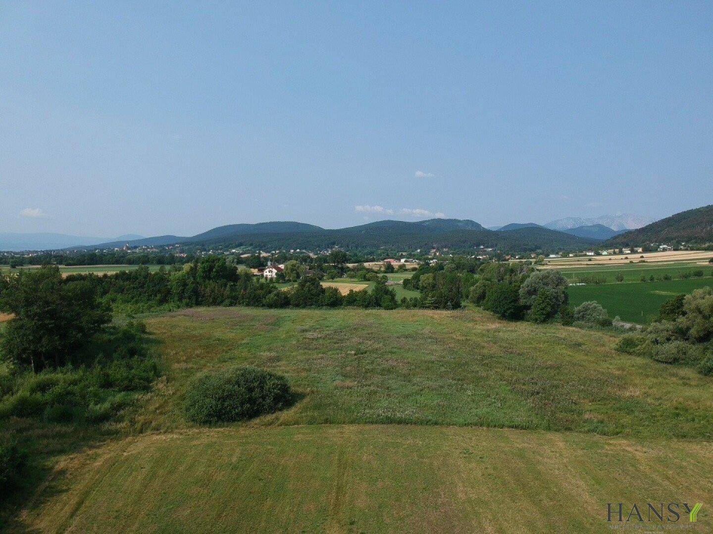 Eine weite Landschaftsaufnahme zeigt grüne Felder, Wälder und Hügel mit vereinzelten Häusern unter blauem Himmel.