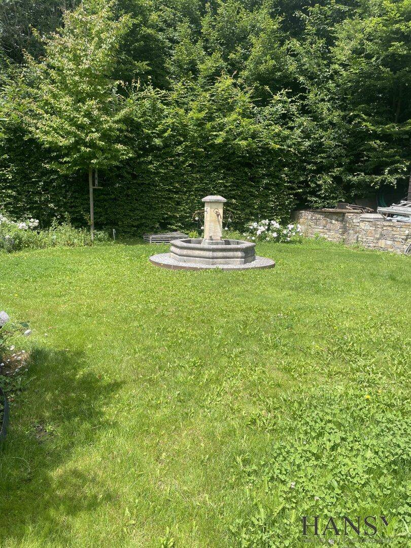 Gepflegter Rasenbereich mit einem dekorativen Steinbrunnen, umgeben von hohen Hecken und Bäumen.