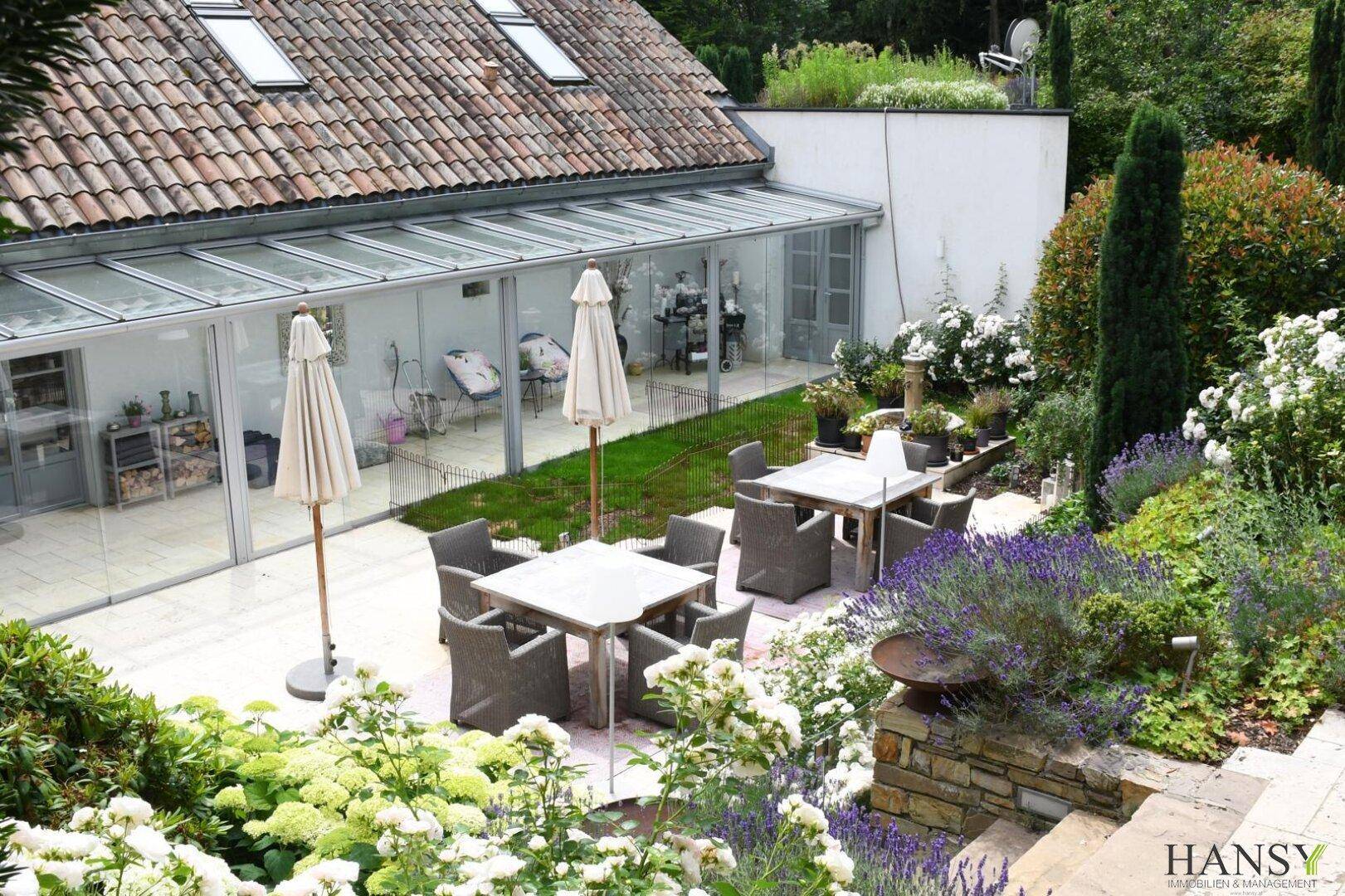 Gemütlicher Essbereich im Freien mit Tischen und Stühlen, umgeben von blühenden Pflanzen und einem Wintergarten.