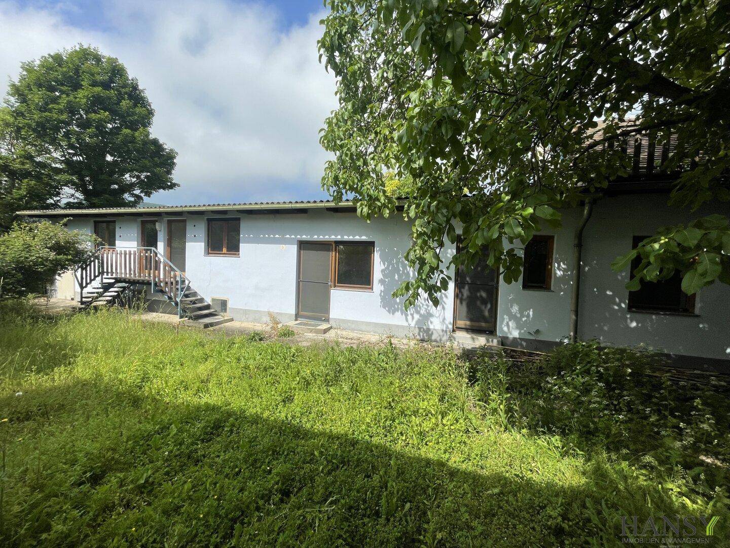 Einfaches Gebäude mit Außentreppe und verwildertem Gartenbereich im Vordergrund.