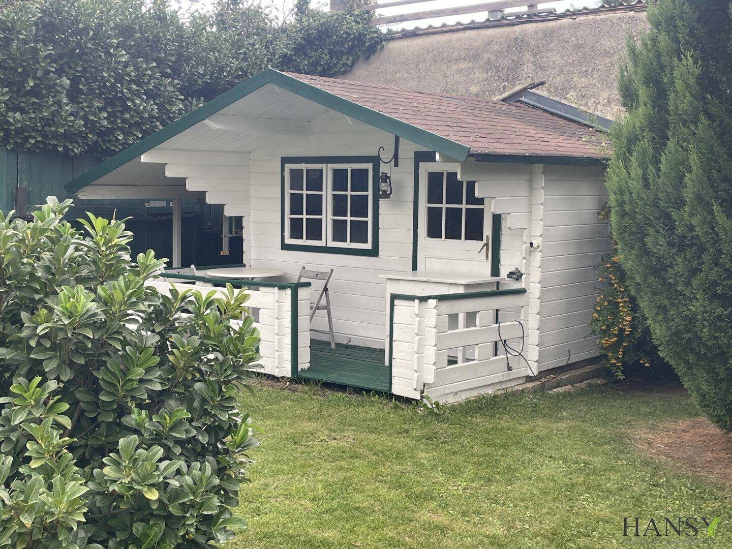 Charmantes weißes Gartenhaus mit grünen Akzenten und einer kleinen Veranda, umgeben von Grün.