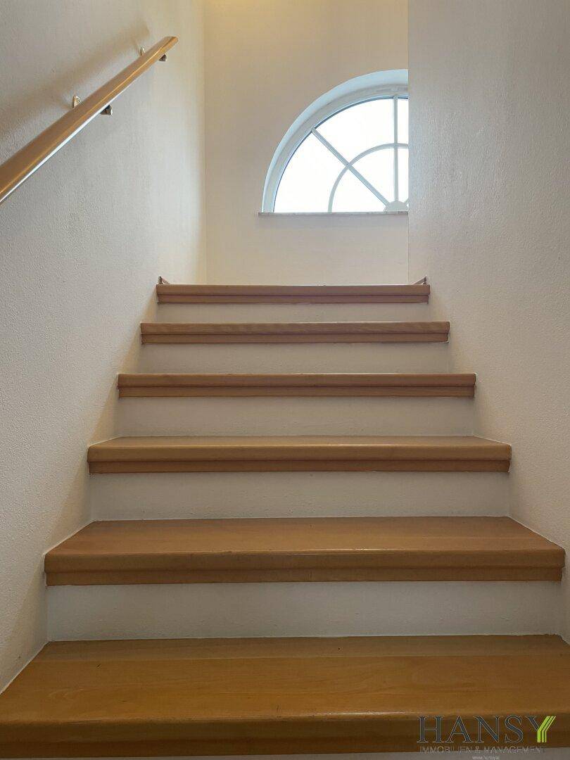 Helle Holztreppe führt in die obere Etage, beleuchtet durch ein dekoratives Rundbogenfenster.