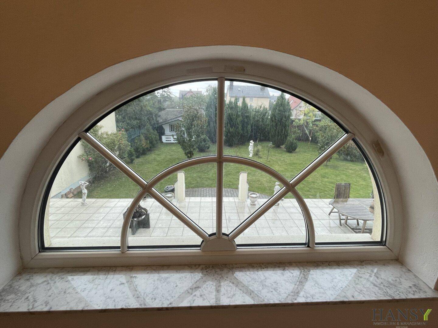 Dekoratives Rundbogenfenster mit Blick auf die Terrasse und den grünen Gartenbereich.