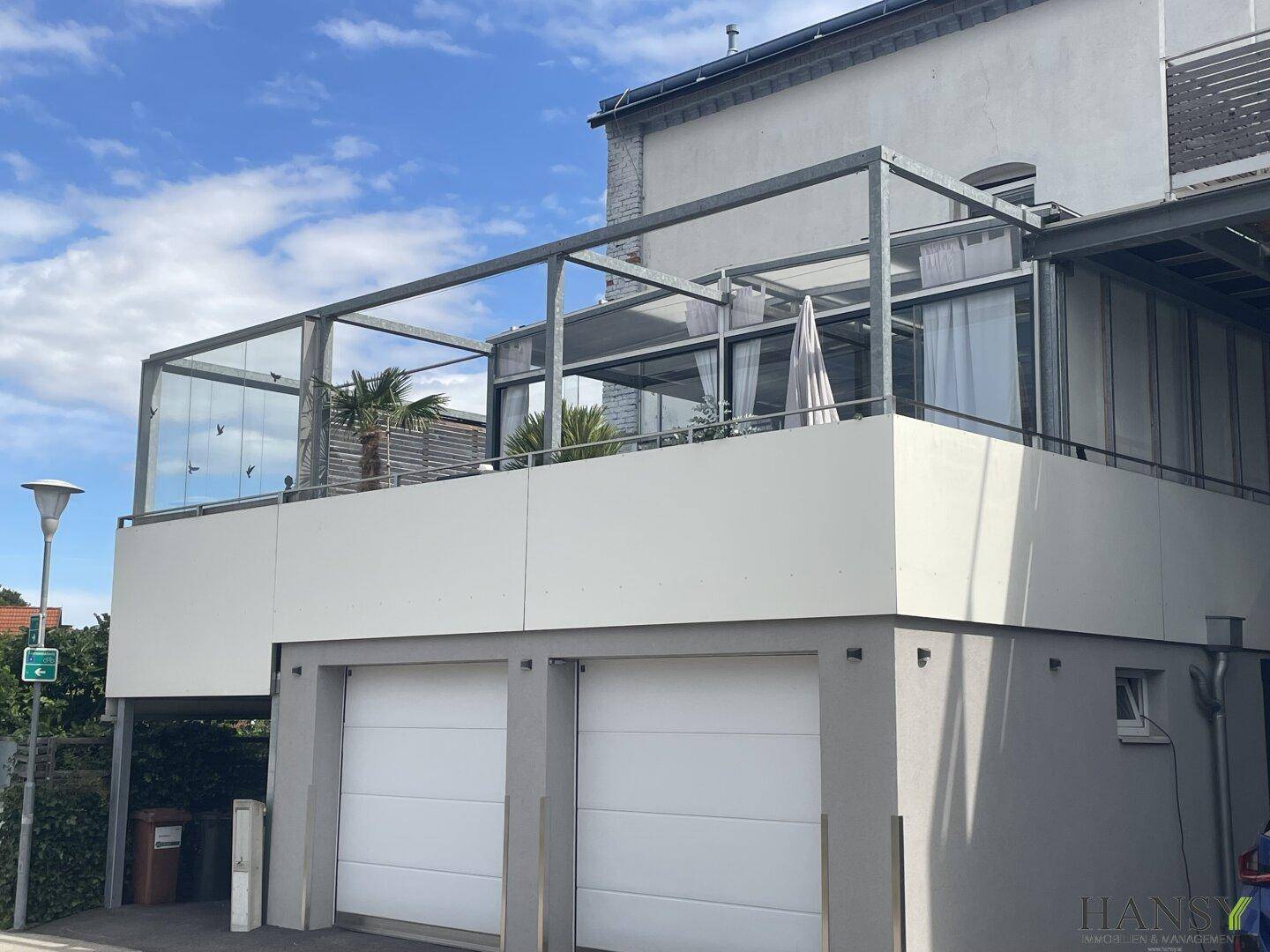 Große moderne Terrasse mit Glasgeländer und Metallkonstruktion über Garagen, geschmückt mit Palmen.