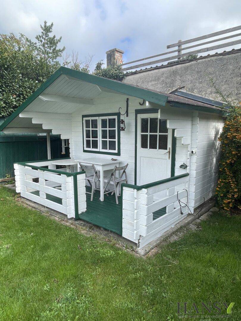 Weißes Gartenhaus mit kleiner Terrasse und Sitzgelegenheiten im Grünen.