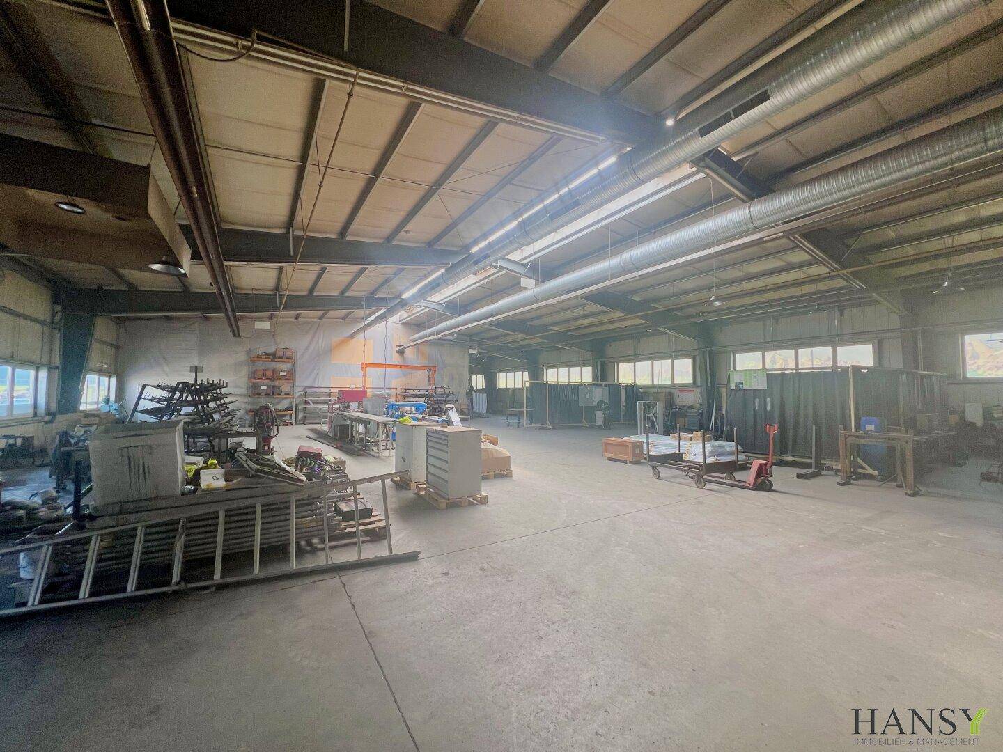 Große Werkshalle mit Betonboden, hohen Decken und sichtbaren Lüftungsrohren, ausgestattet für Produktion und Montage.