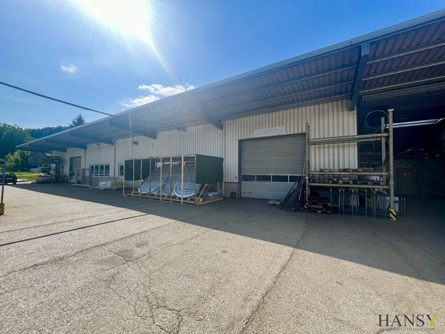 Industrielle Fassade mit Rolltor und Vordach, umgeben von einem großen asphaltierten Parkplatz unter strahlendem Himmel.