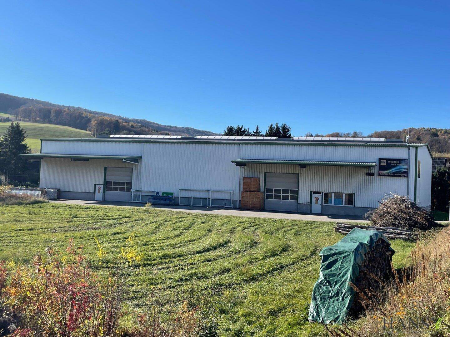 Industrielles Gebäude mit Photovoltaikanlage auf dem Dach, umgeben von einer weiten Grünfläche unter blauem Himmel.