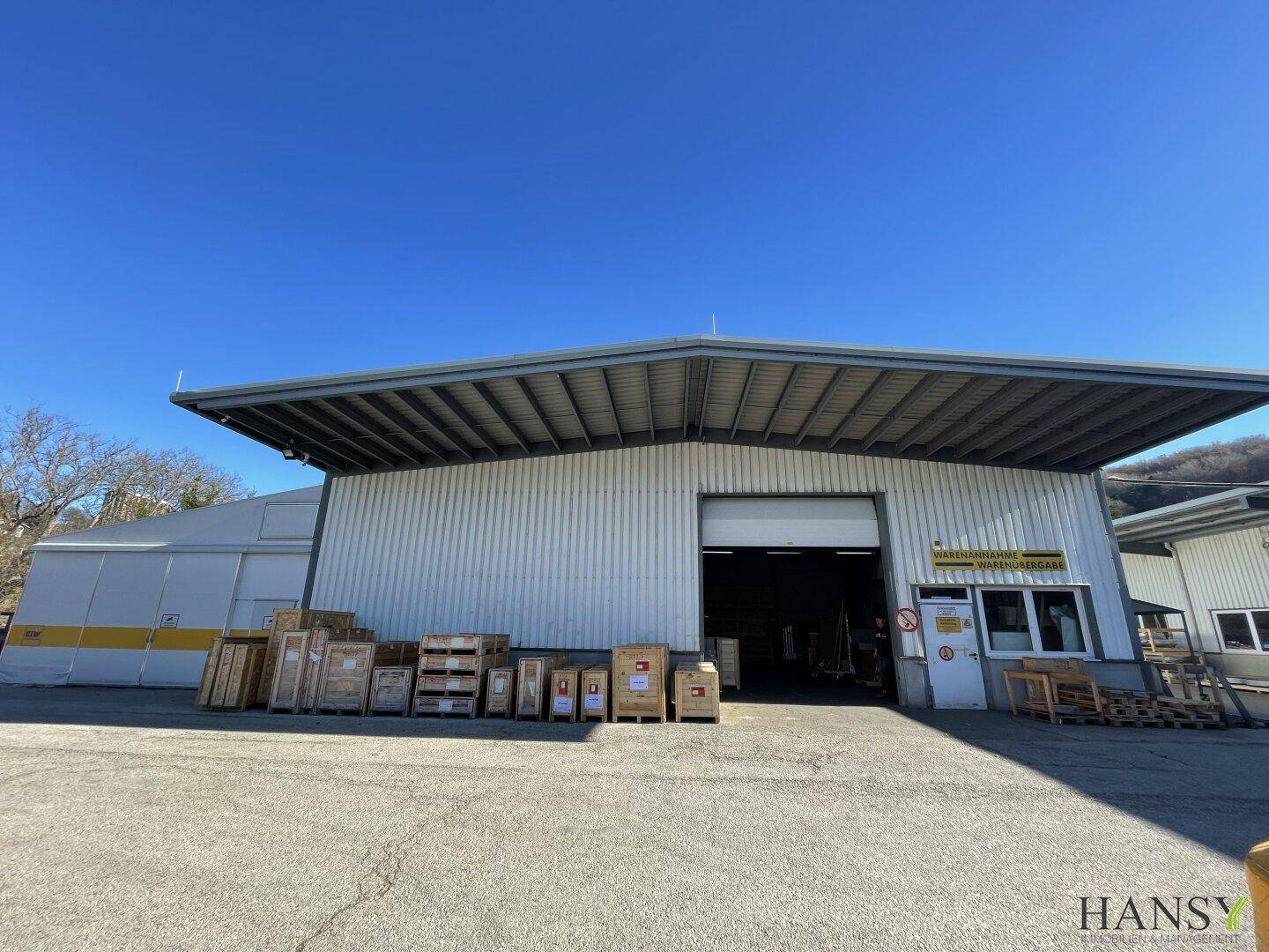 Große Lagerhalle mit offenem Rolltor und Vordach, umgeben von einem weiten asphaltierten Bereich unter klarem Himmel.