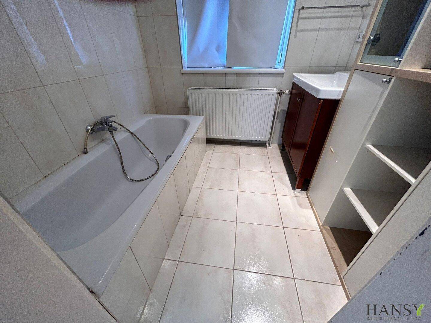 Helles Badezimmer mit Badewanne, Waschbecken und Fliesenboden, ausgestattet mit einem Heizkörper unter dem Fenster.