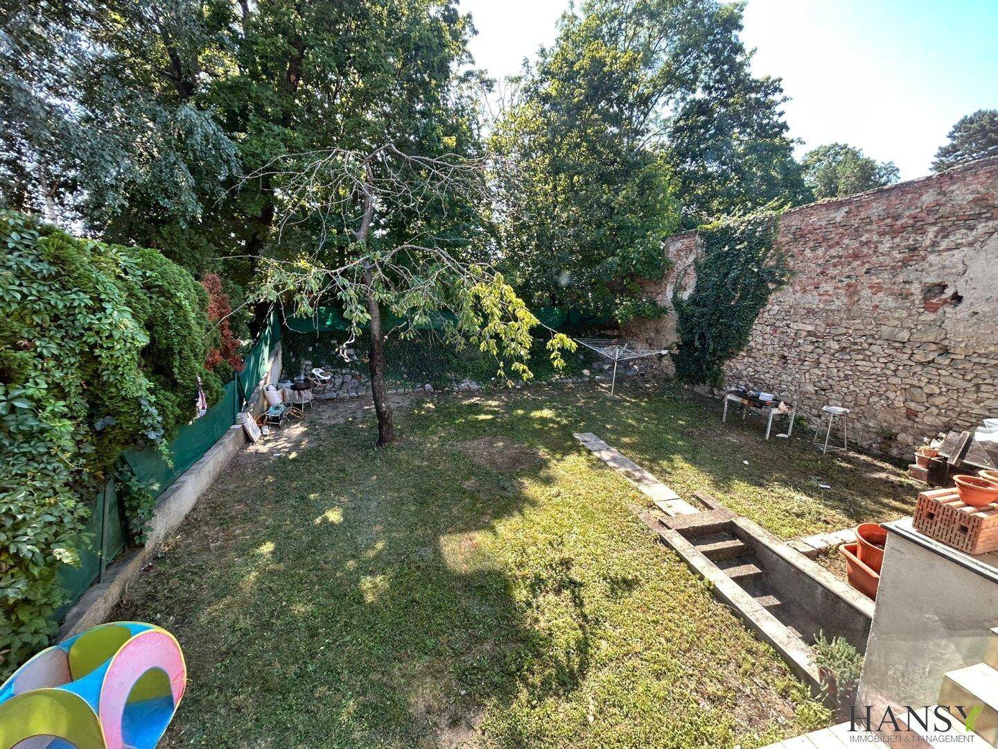 Grüner Gartenbereich mit Rasenfläche, Bäumen und einer historischen Steinmauer im Hintergrund.