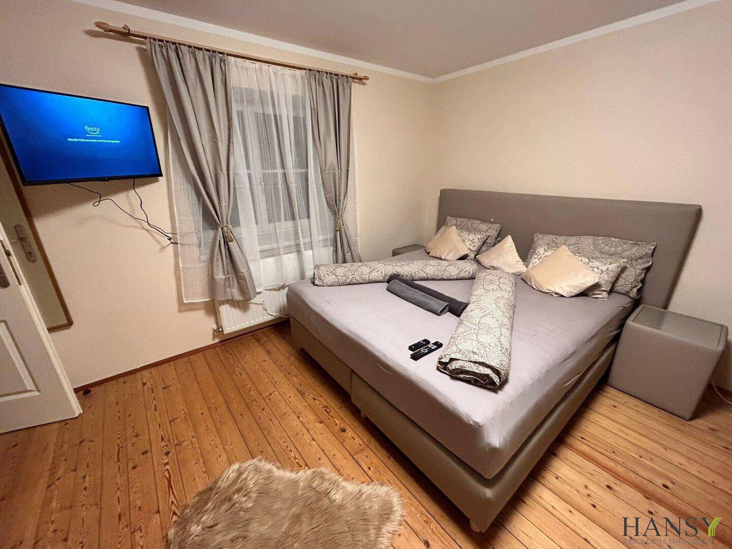 Schlafzimmer mit bequemem Doppelbett, an der Wand montiertem Fernseher und Holzboden.