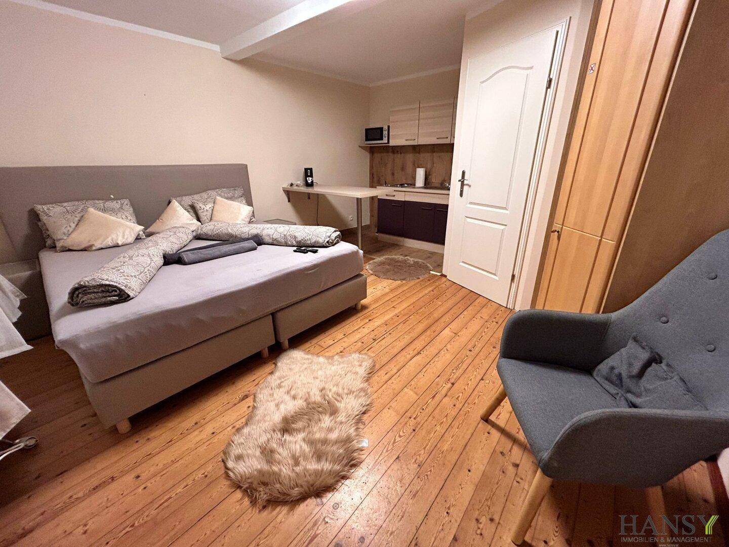 Kombiniertes Schlafzimmer mit Bett, Sessel und integrierter kleiner Küchenzeile.