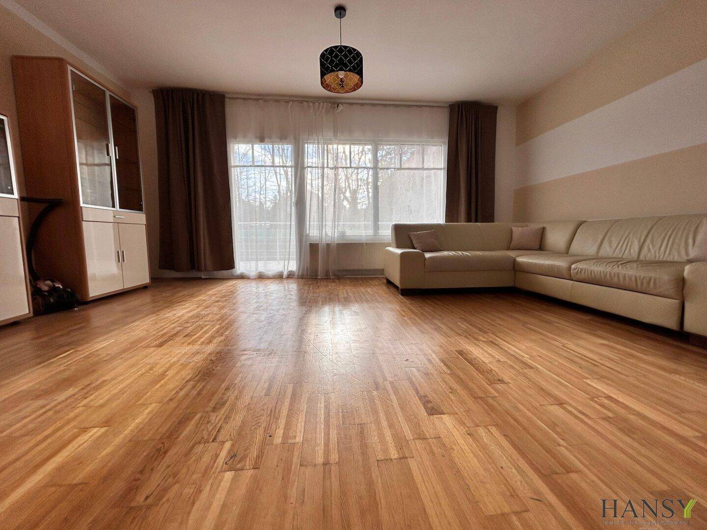 Helles Wohnzimmer mit beigem Sofa, Schränken und großen Fenstern, die viel Licht hereinlassen.